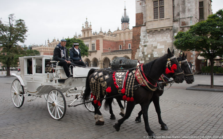 White horse-drawn carriage with two decorated black horses crossing Kraków’s Main Market Square.