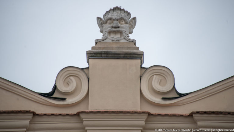 Carved grotesque head with pointed ears above a pale curving historic gable in Kraków.
