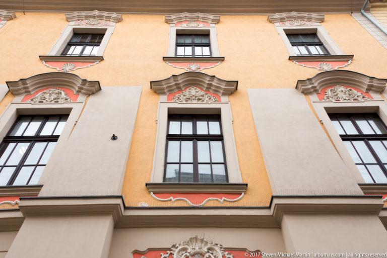 Pastel Kraków townhouse façade with tall windows, red-and-cream ornamental panels, scrolls, and molded cornices.