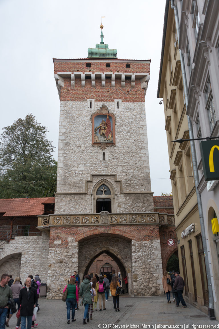 St. Florian’s Gate in Kraków, with rough stone masonry, pointed passage, crenellations, religious image, and visitors below.