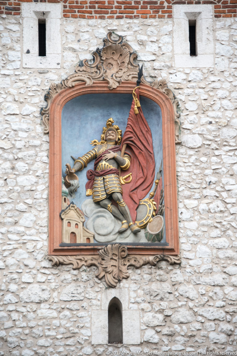 Painted relief of St. Florian in armor with banner and water vessel, mounted on the stone wall of St. Florian’s Gate.