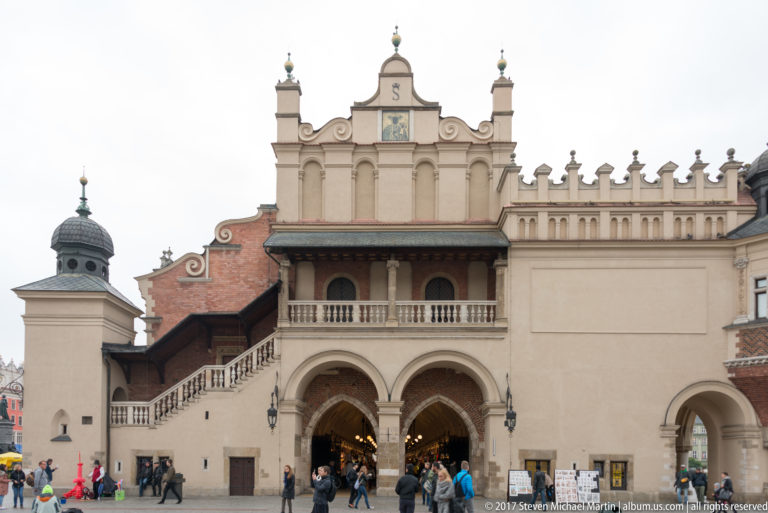 Historic Kraków gateway with twin arches, balcony, ornate stepped attic, finials, and pedestrians passing beneath.