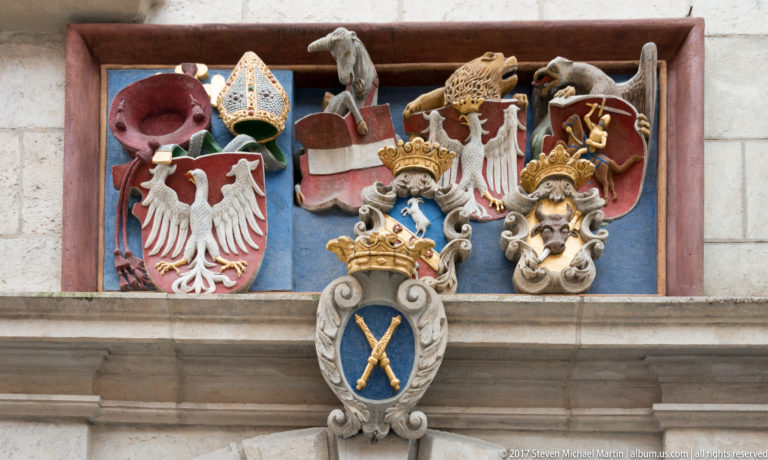 Group of carved and painted coats of arms on a Kraków façade, including crowned white Polish eagles and other heraldic emblems.