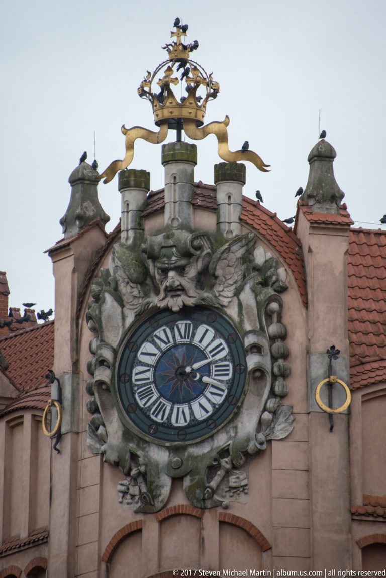 Large Roman-numeral clock in an ornate sculpted surround on a historic Kraków façade, topped by gilded heraldic ornament.