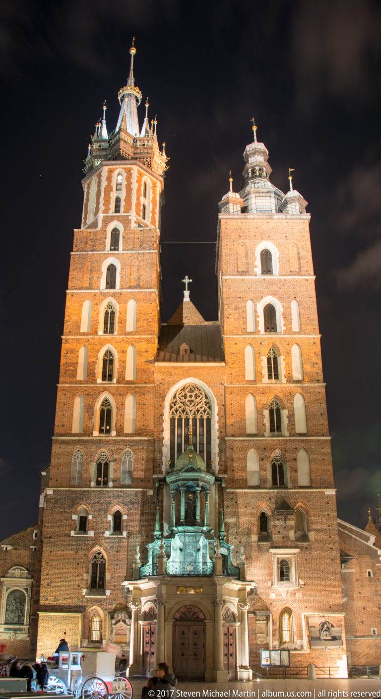 Closer night view of St. Mary’s Basilica with its ornate taller tower, shorter companion tower, and traceried Gothic window.