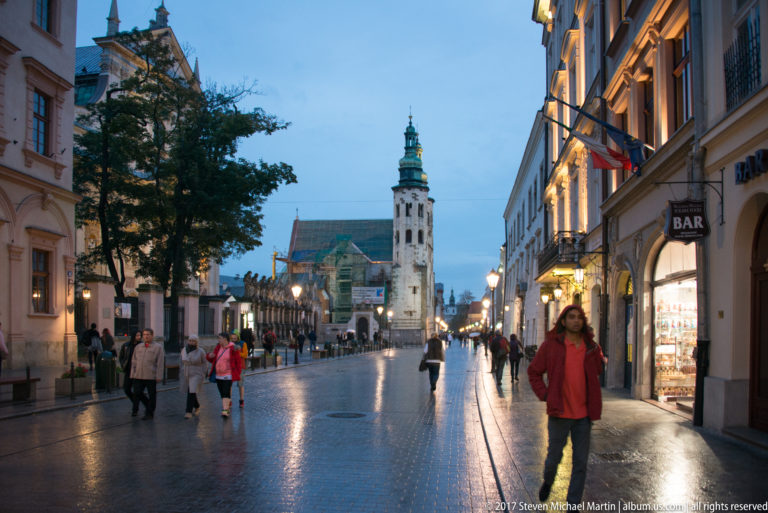 Rain-wet pedestrian street in Kraków at dusk, with historic façades, pedestrians, shop lights, and a green-capped church tower ahead.