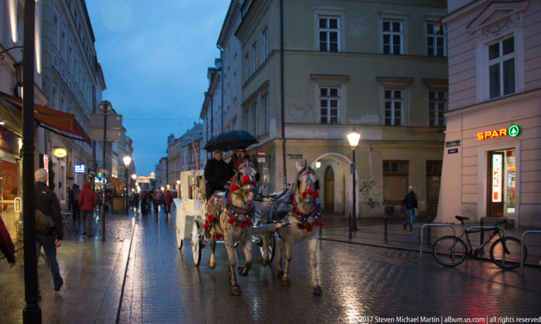 Two decorated white horses pulling a carriage along a rain-wet Kraków street at dusk, with pedestrians and shops nearby.