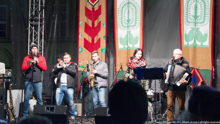 Outdoor band in Kraków performing with trumpet, saxophone, woodwind, and accordion before colorful folk-patterned textile panels.