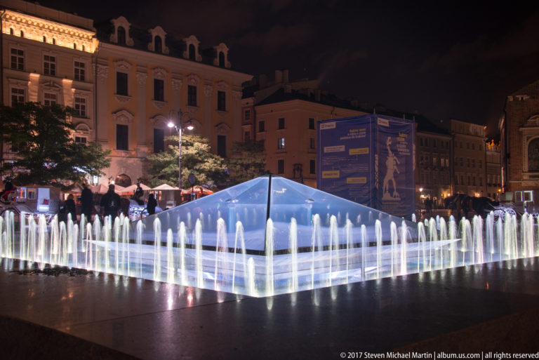 Illuminated geometric fountain with rows of white water jets in a Krakow square, surrounded by historic buildings at night.