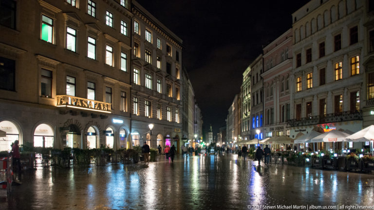 Rain-wet Kraków Old Town street at night with illuminated historic façades, cafés, umbrellas, and reflections on the paving.