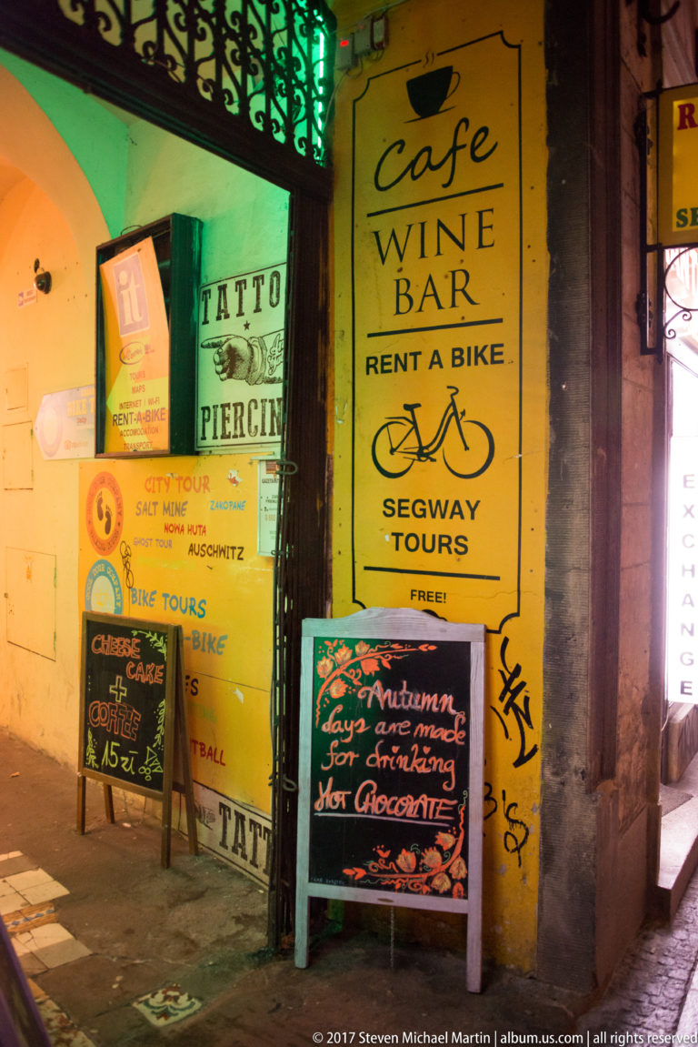 Nighttime Kraków storefront crowded with signs for a café, wine bar, bike rental, Segway tours, tattooing, piercing, and hot chocolate.