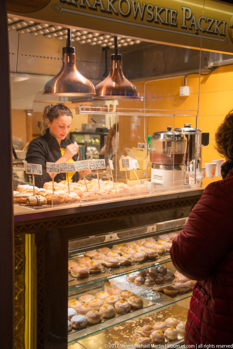 Krakowskie Pączki shop counter in Kraków with pastries behind glass, handwritten flavor labels, copper lamps, seller, and customer.