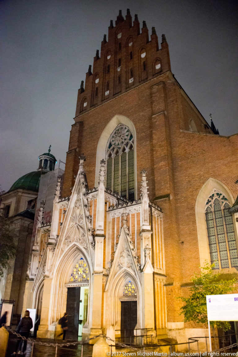 Brick Gothic façade of Kraków’s Dominican Basilica of the Holy Trinity with traceried window, two pointed portals, and illuminated stone details.