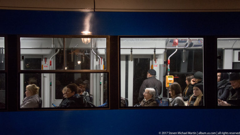 Passengers seated and standing inside a blue Kraków tram, visible through large windows with handrails and ticket equipment.