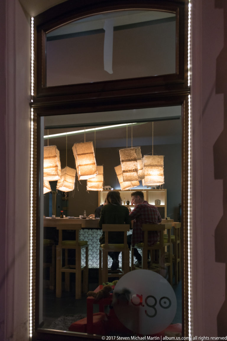 Modern Kraków restaurant interior seen through a lit window, with diners at wooden tables beneath large rectangular pendant lamps.