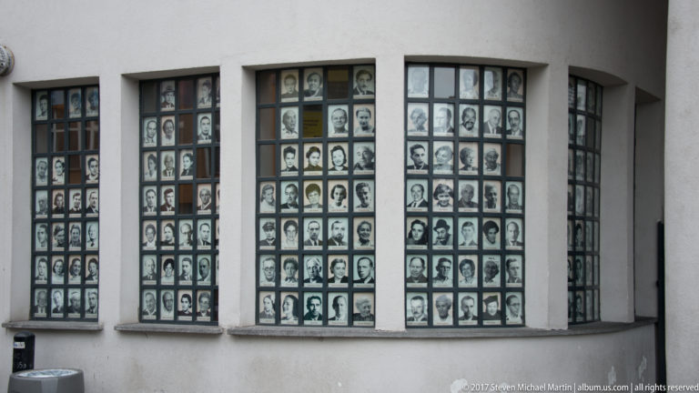 Ground-floor windows of Oskar Schindler’s former factory in Kraków filled with grids of black-and-white portrait photographs.