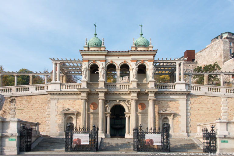 Front view of the Castle Garden Bazaar entrance pavilion with twin green domes, arcade, sculptures, and brick wings.