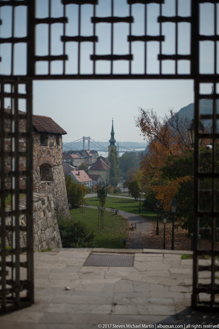 View through a dark metal gate from Castle Hill toward lower Buda, rooftops, and a distant bridge.