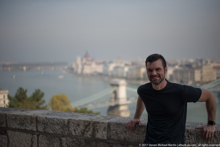 Man leaning on a stone parapet with the Chain Bridge, Danube, and Pest skyline blurred behind him.