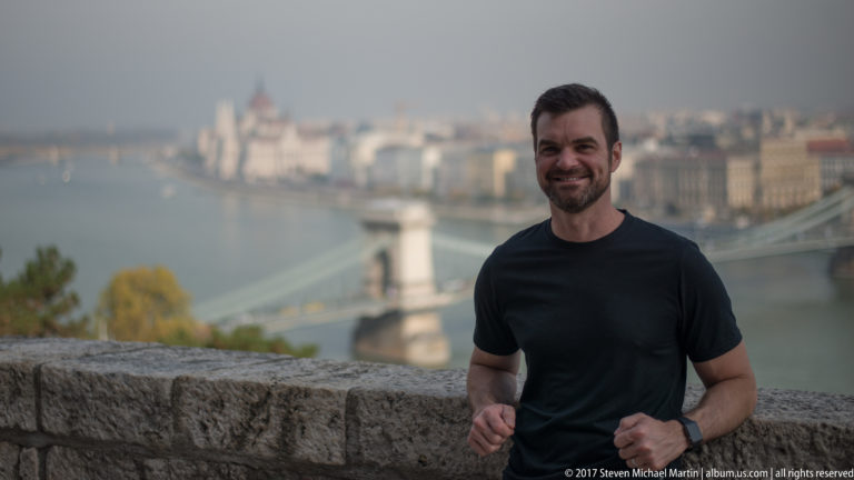 Man standing by a stone parapet with the Danube and Chain Bridge softly blurred in the background.