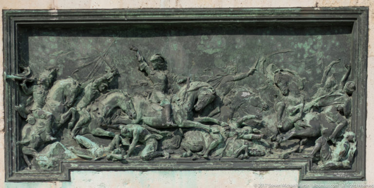 Bronze battle relief with cavalry, fallen soldiers, weapons, and horses on the Prince Eugene monument pedestal.