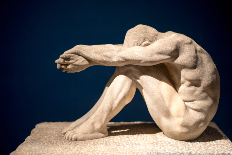 Seated nude male sculpture with knees raised and both arms extended forward across the face.