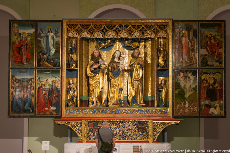 Large open winged altarpiece with three central saints, carved figures, gilding, and multiple painted narrative panels.