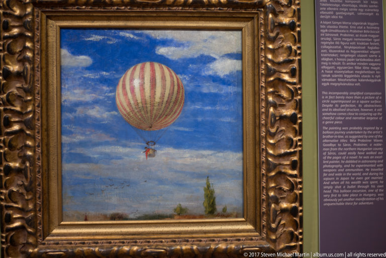 Small framed painting of a red-and-white striped hot-air balloon floating in a blue, clouded sky.