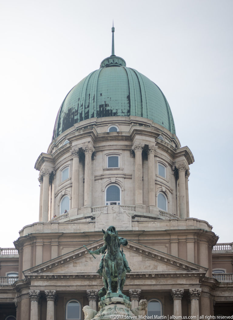 Green dome of the Royal Palace above a columned classical façade and bronze rooftop sculpture.