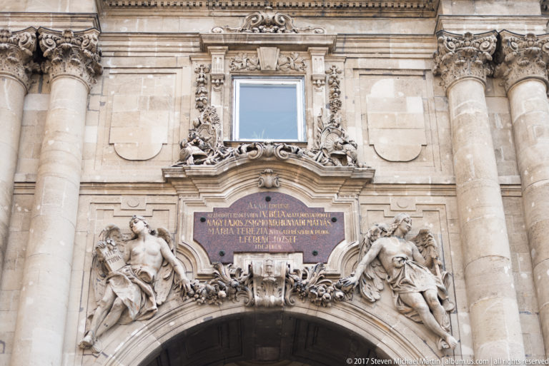 Ornate Baroque arched portal with Latin inscription, reclining sculpted figures, columns, and elaborate stone ornament.