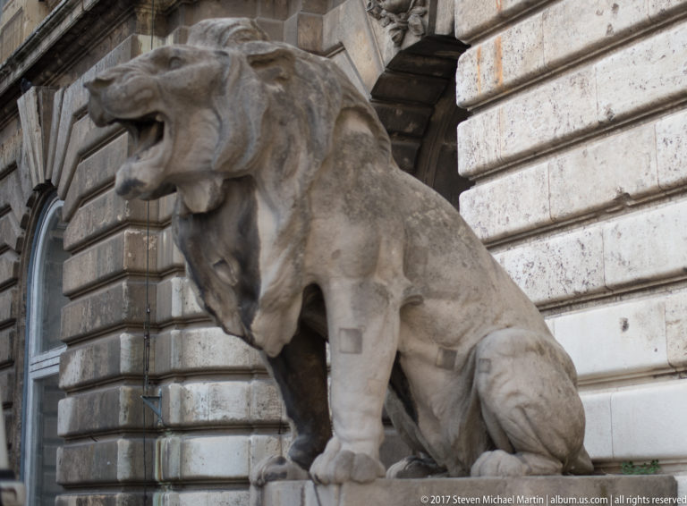Large seated stone lion with open mouth beside a rusticated stone wall at Buda Castle.