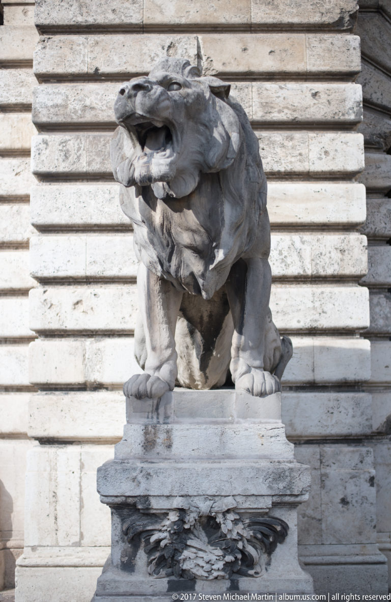 Front view of a crouching stone lion with open mouth on an ornamental pedestal at Buda Castle.