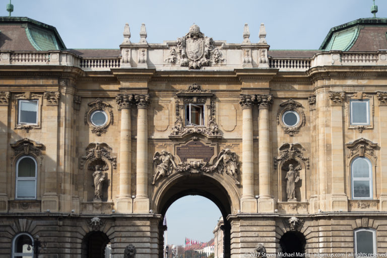 Large arched palace gateway with paired columns, statues, heraldic ornament, and an inscription panel.