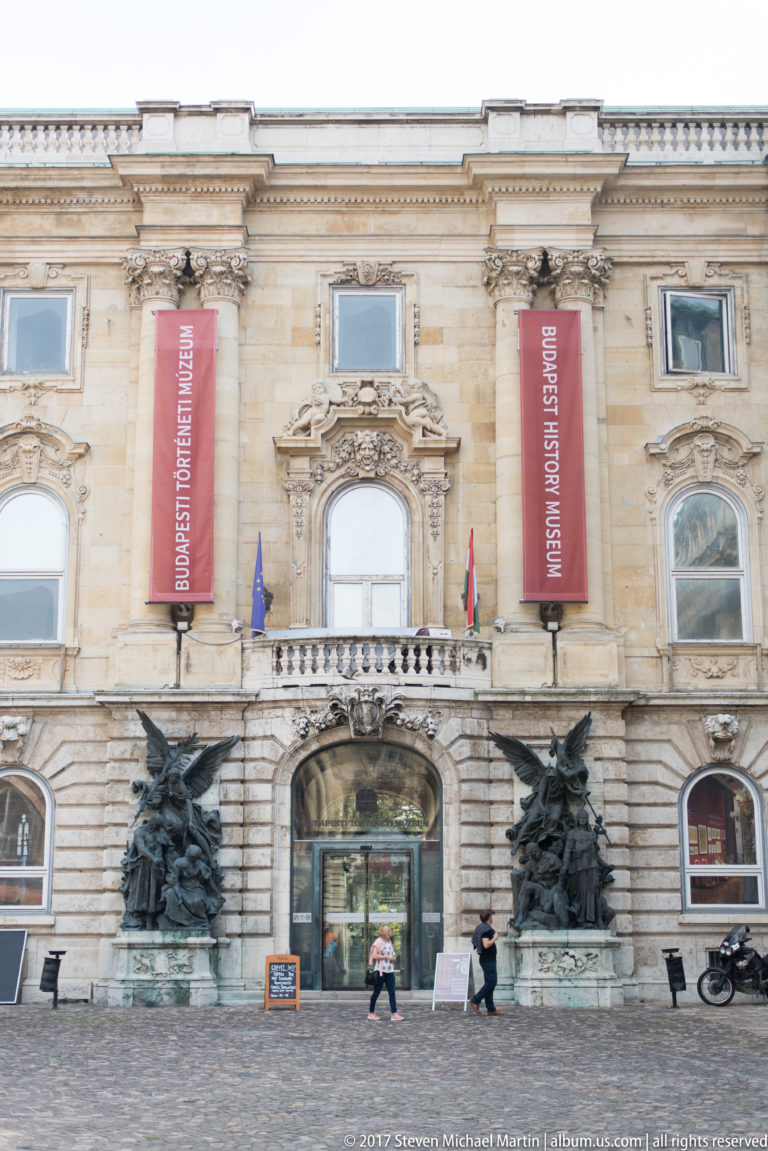 Budapest History Museum entrance with red banners, flags, bronze sculptures, and pale stone palace façade.