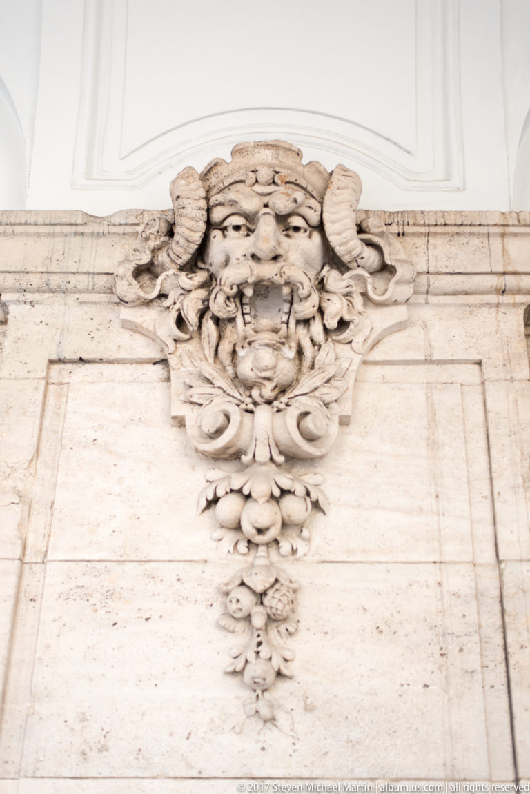 Carved grotesque face with curled forms and hanging leaf ornament on pale stone palace architecture.