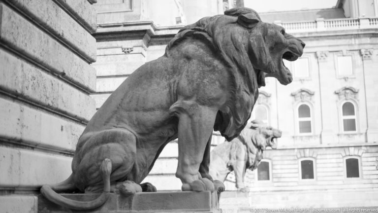 Profile of a large stone lion with open mouth beside the palace wall, with another lion in the distance.