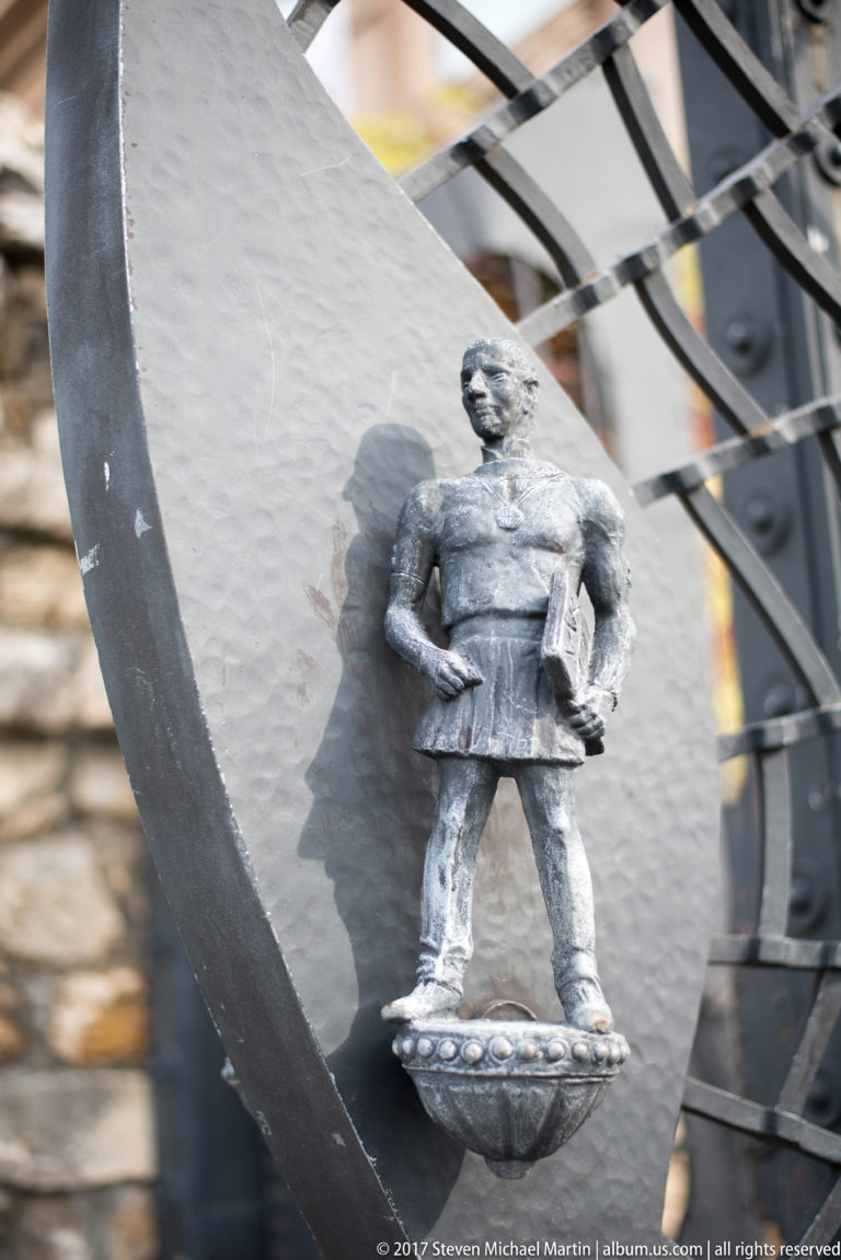 Small cast-metal soldier figure standing on an ornamental bracket attached to a black iron gate.