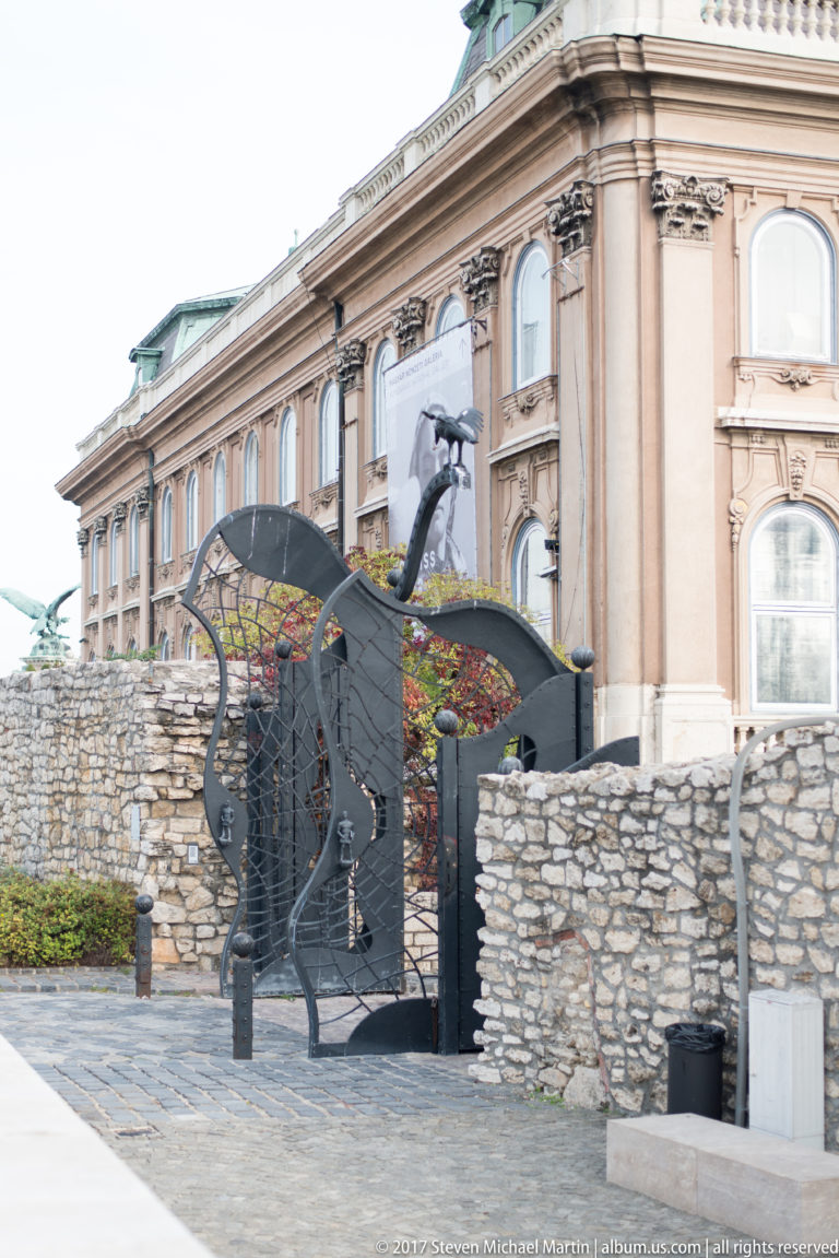 Curving black wrought-iron gate between low stone walls with the palace façade rising behind.