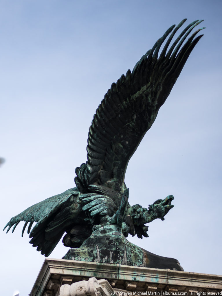 Bronze Turul bird with wings spread wide on a monument at Buda Castle, silhouetted against the sky.