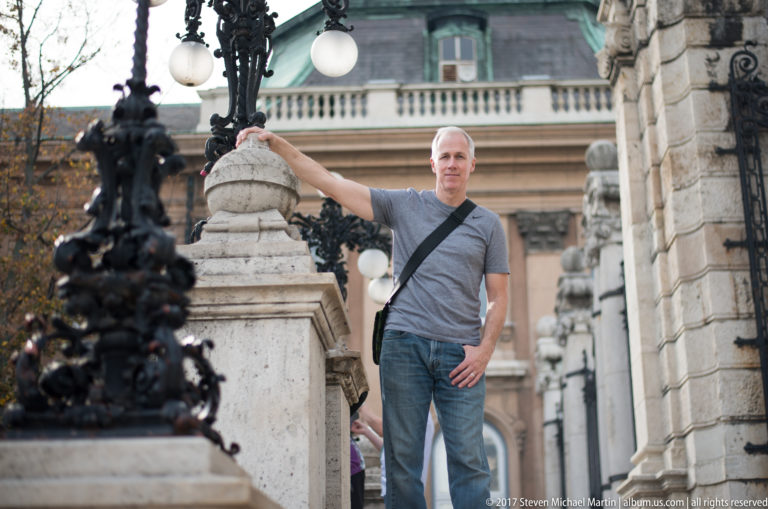 Man in a gray T-shirt posing beside a stone balustrade and ornate black lamp at Buda Castle.
