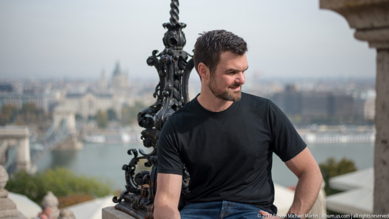 Bearded man in a black shirt seated at Castle Hill with the Danube and Pest skyline blurred behind.