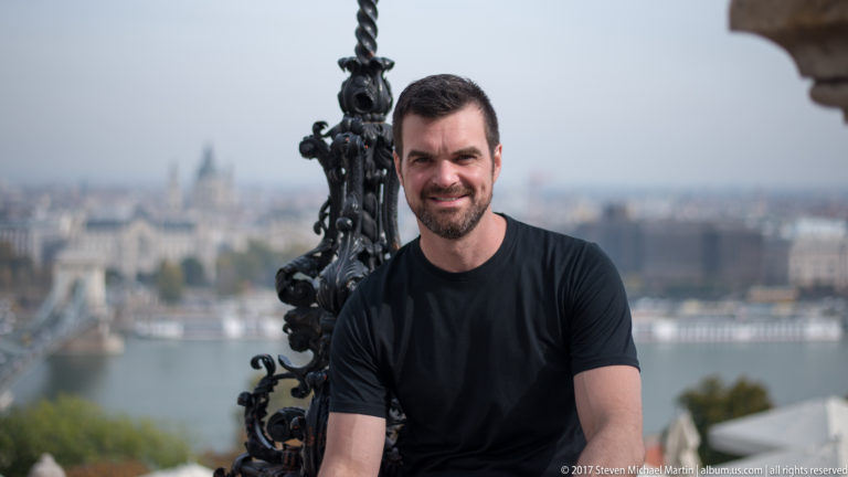 Bearded man smiling at the camera with the Danube and Budapest skyline softly blurred behind.