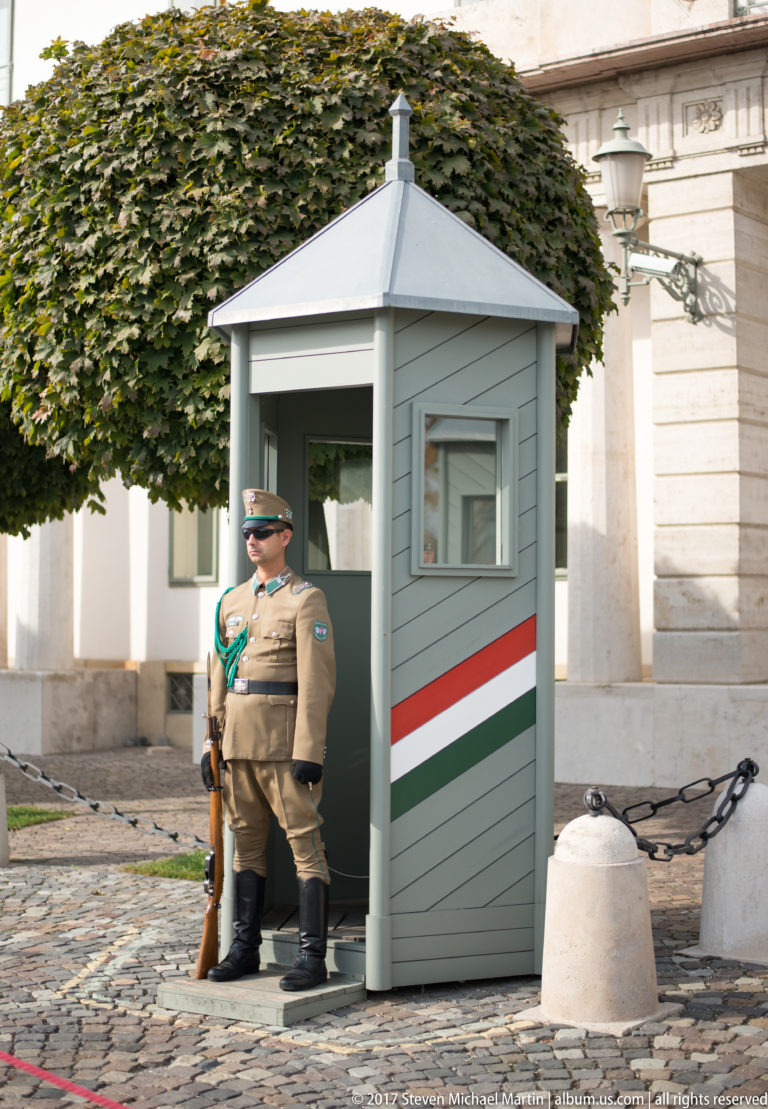 Hungarian ceremonial guard holding a rifle beside a gray sentry box marked with red, white, and green stripes.