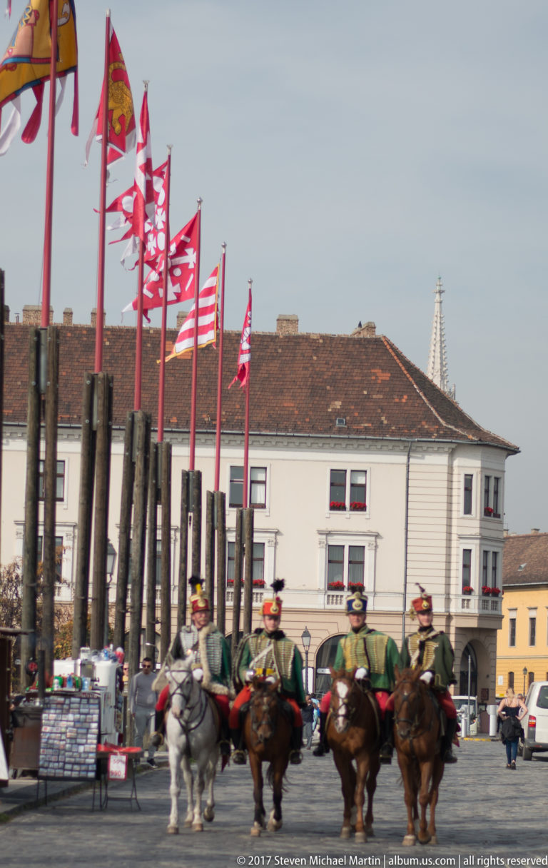 Mounted ceremonial guards in historic-style uniforms riding across a paved square lined with red-and-white flags.