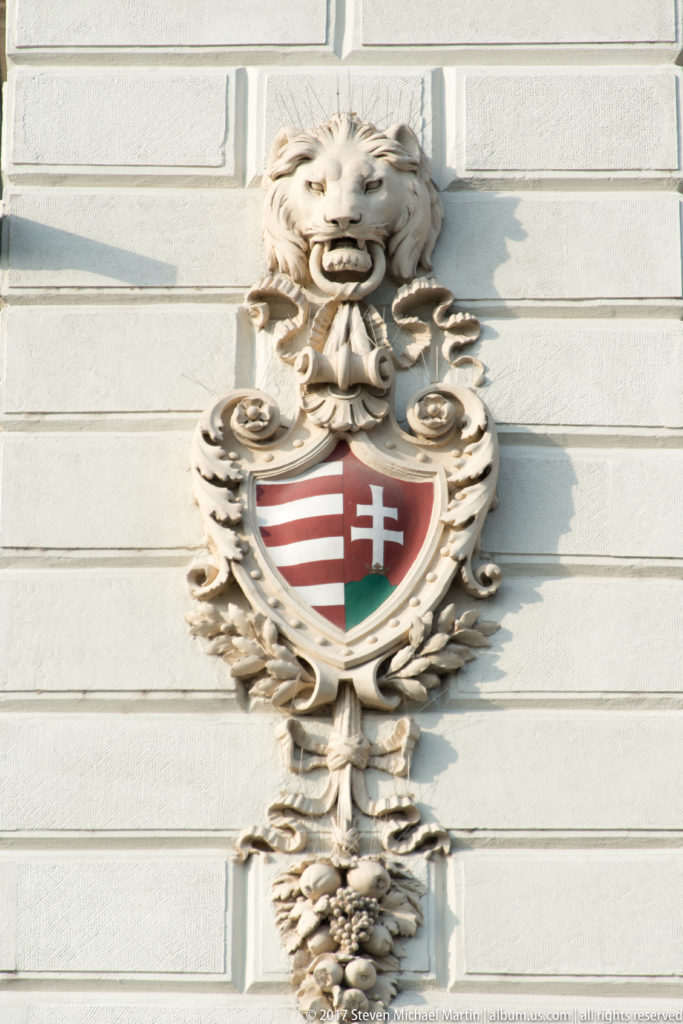 Crowned Hungarian coat of arms in sculpted scrollwork beneath a lion’s head on a pale stone façade.