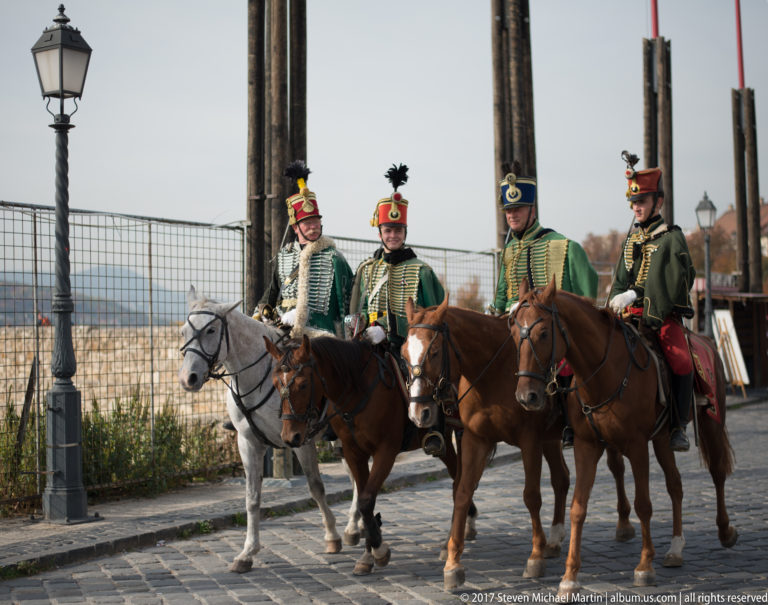 Four mounted ceremonial guards in plumed historic-style uniforms riding side by side on gray and brown horses.