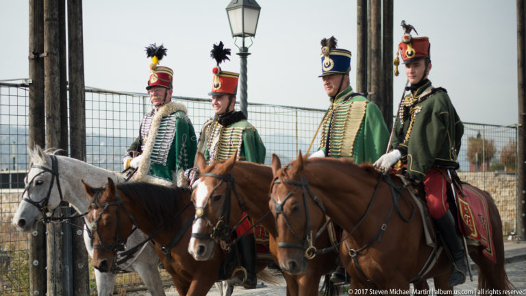 Four mounted ceremonial guards in green braided uniforms and tall plumed headgear ride gray and brown horses.