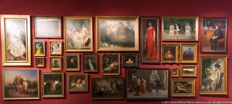 Dense salon-style display of framed portraits, landscapes, interiors, and figure paintings on a deep red museum wall.