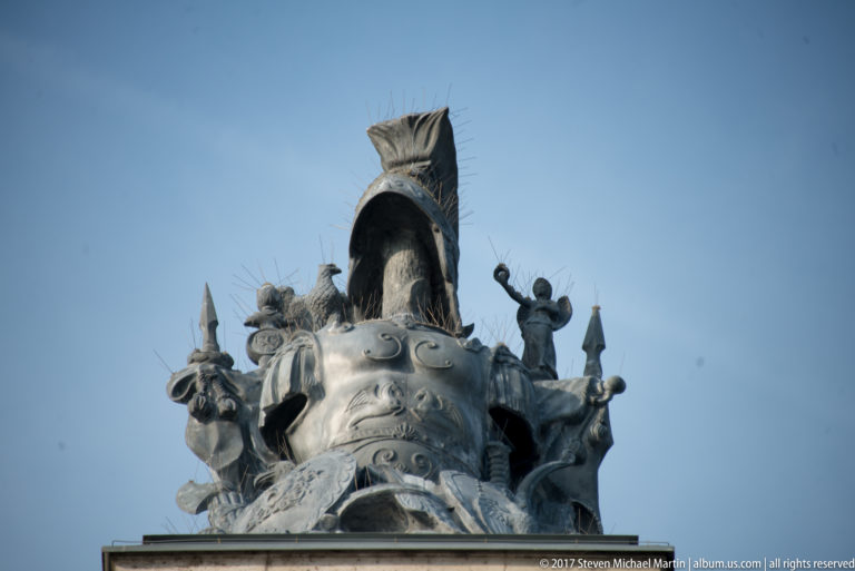 Metal rooftop sculpture with a large crested helmet, small figures, and weapon motifs against a blue sky.