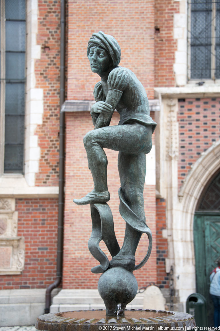 Bronze seated figure on an abstract curling support, with the red-brick wall and Gothic opening of St. Mary’s Basilica behind.